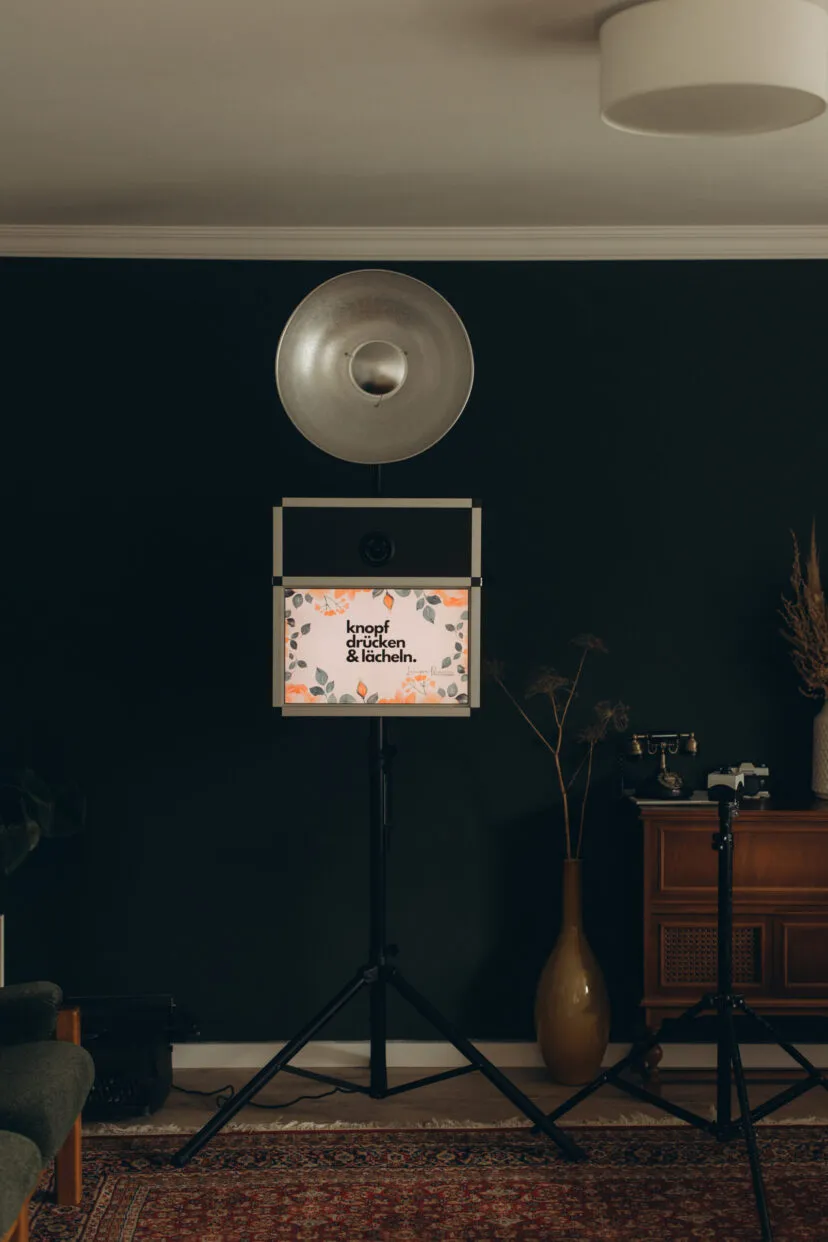 Fotobox mit Blitz-Reflektor und Bildschirm „knopf drücken & lächeln.“ auf einem Stativ in einem Wohnzimmer